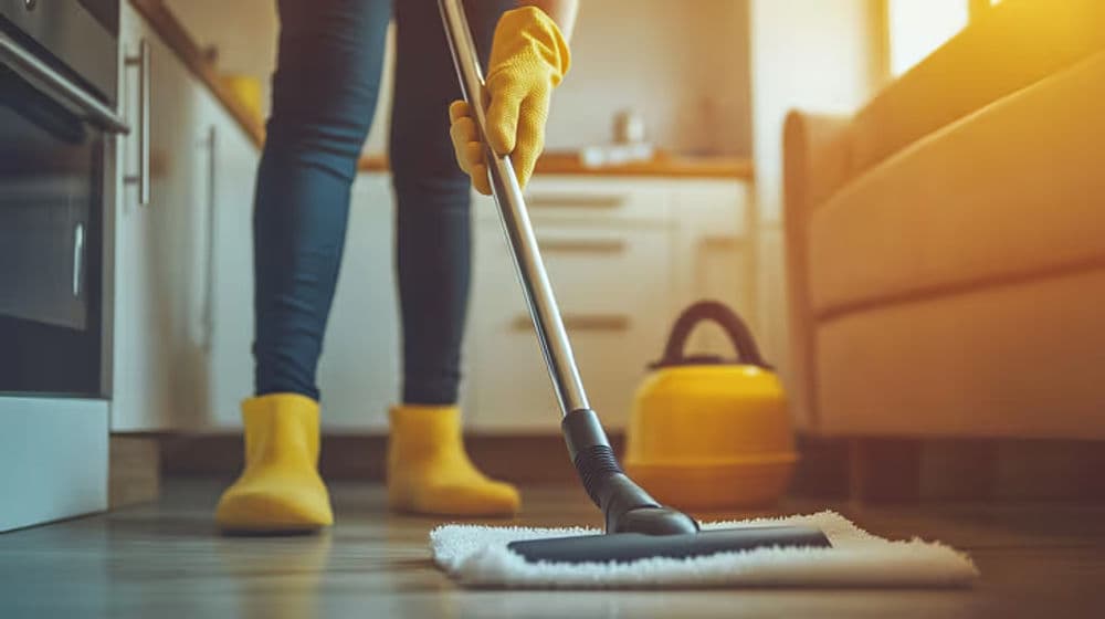 Person wearing yellow gloves mopping a kitchen floor with a vacuum cleaner in the background.